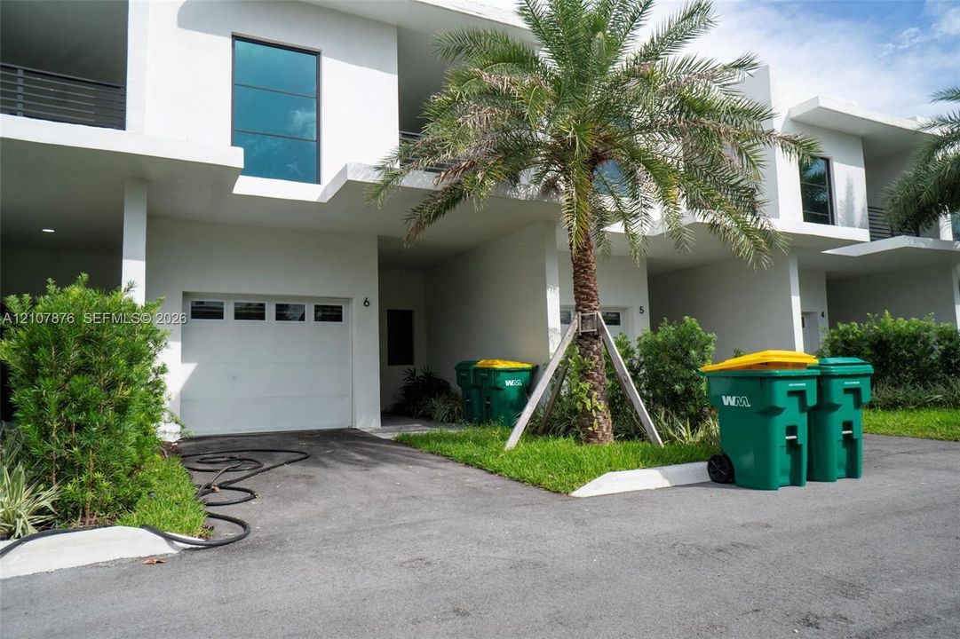 Modern townhouse exterior featuring white stucco siding, integrated single-car garages, second-story balconies with glass railings, and mature palm landscaping