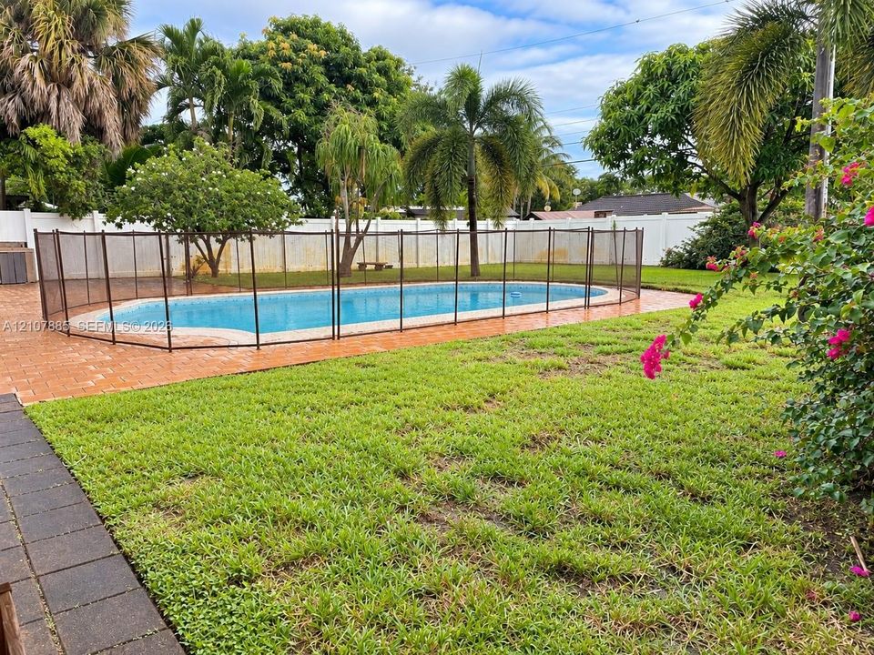 Backyard featuring an in-ground swimming pool, mesh safety fencing, paved patio, expansive lawn, and white perimeter privacy fencing