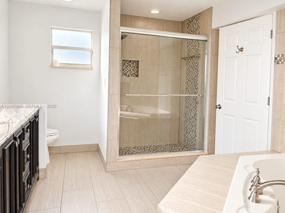 Walk-in shower with sliding glass door, mosaic tile accent wall, built-in shower niche, light-toned floor tile, and dark wood-finish cabinetry with stone countertop