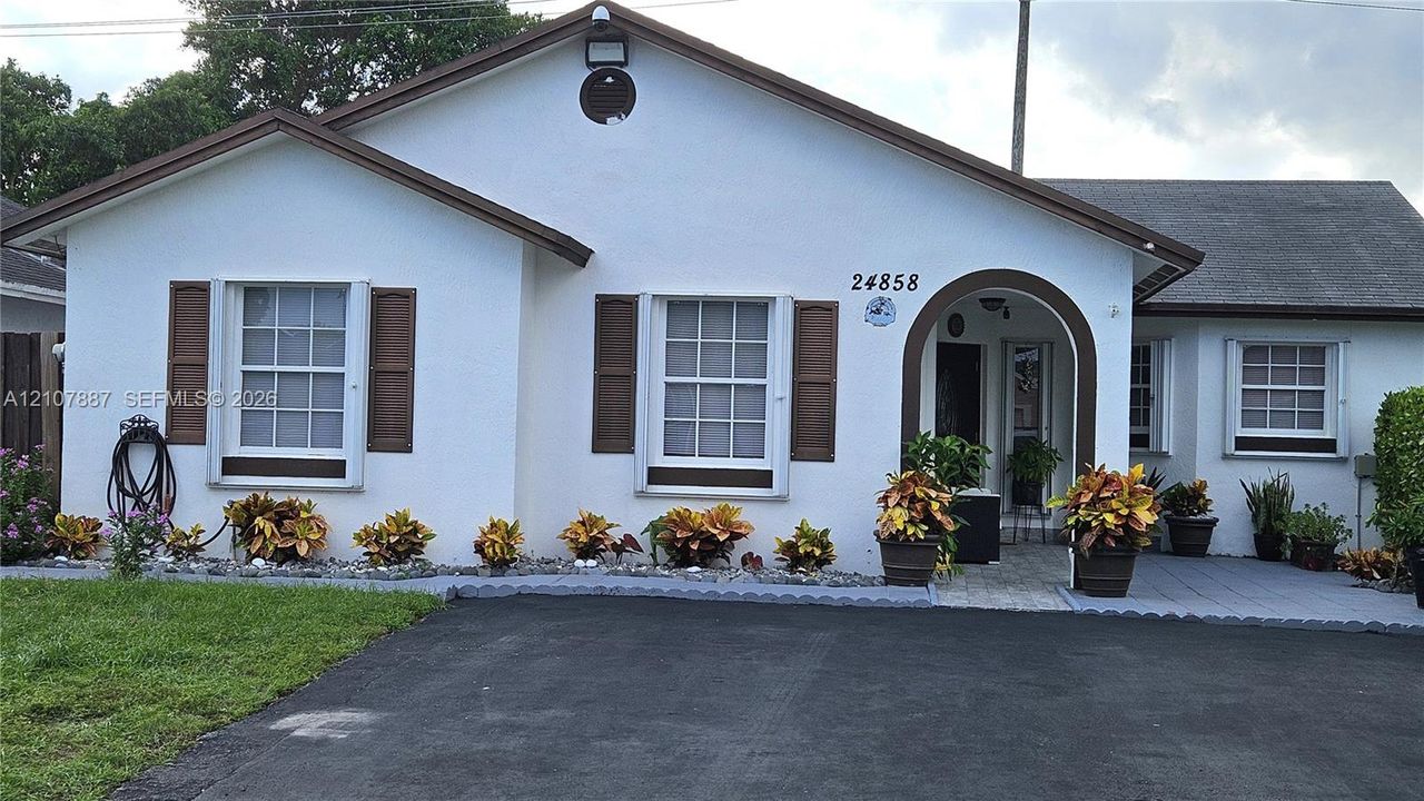 Stucco exterior featuring arched entryway, multi-pane windows with decorative and accordion shutters, shingled roof, and paved driveway