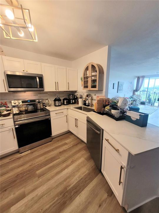 Kitchen featuring shaker-style cabinetry with brass-finish pulls, quartz countertops, stainless steel appliances, a geometric chandelier, and wood-finish flooring