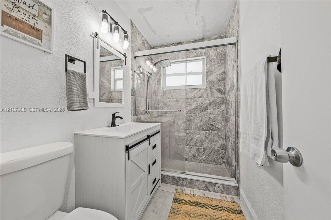 Bathroom featuring a white farmhouse-style vanity with matte black hardware, a rectangular sink with a matching faucet, a tri-light fixture, and a tiled shower enclosure with sliding glass doors