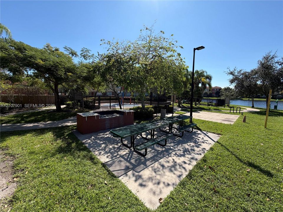 Outdoor recreation space featuring a concrete grilling station, metal picnic tables, paved walkways, and mature landscaping