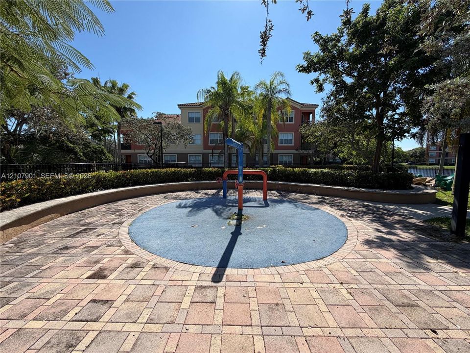 Splash pad featuring a circular rubberized play surface, integrated water feature, paved brick surround, and perimeter landscape hedging