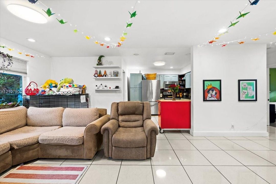 Open-concept interior featuring light-colored tile flooring, recessed lighting, white wall-mounted shelving, and a kitchen equipped with stainless steel appliances and cabinetry