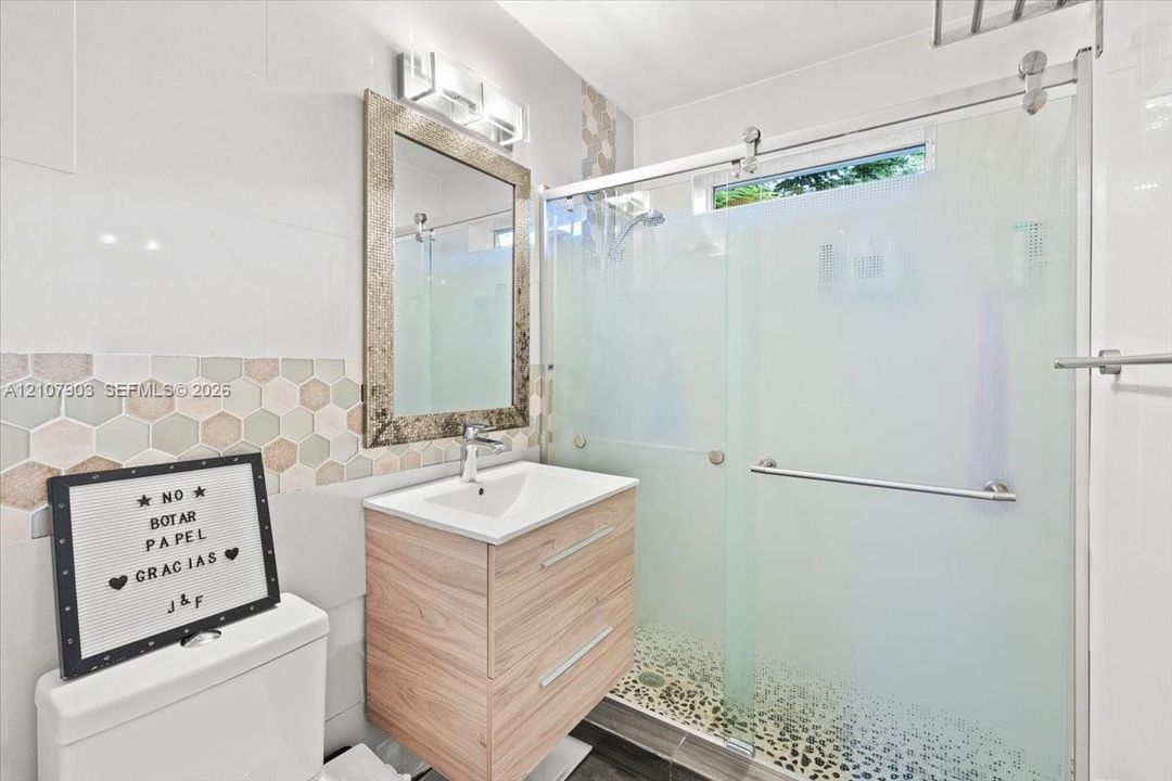 Bathroom featuring a wood-finish vanity with a rectangular sink, walk-in shower with frosted glass sliding doors, stone-tiled shower floor, and hexagonal wall tile backsplash