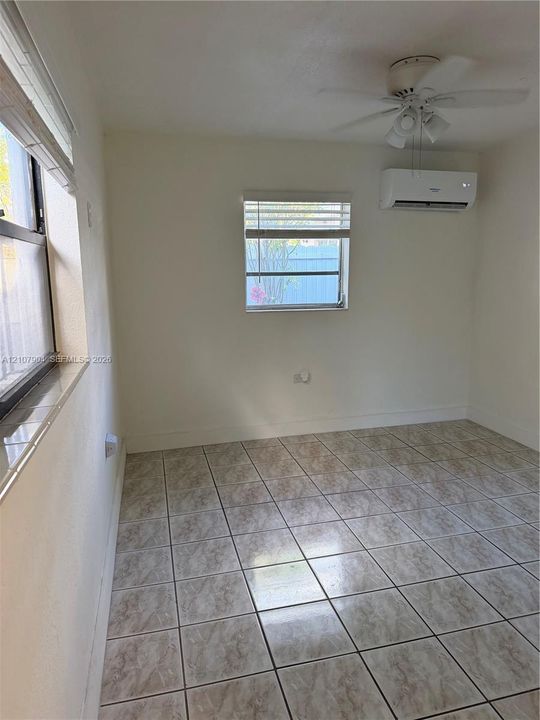 Room featuring stone-finish tile flooring, a wall-mounted ductless air conditioning unit, a multi-blade ceiling fan with an integrated light fixture, and horizontal blind window coverings