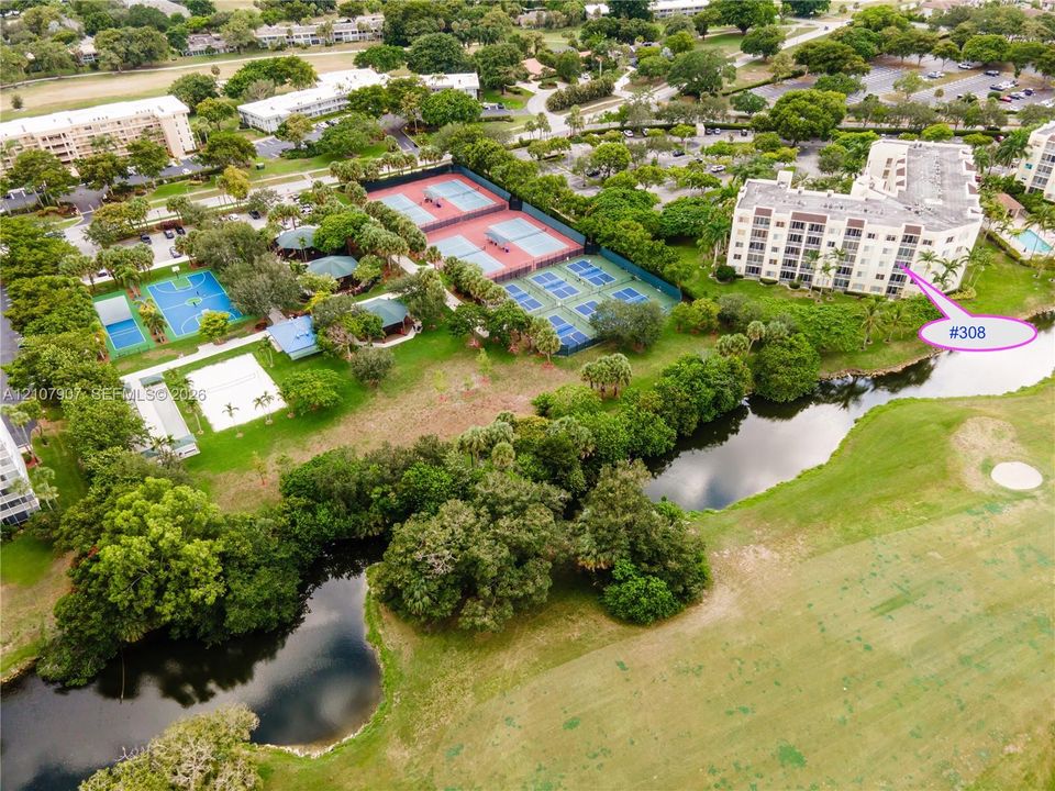 Aerial perspective of a multi-story residential building featuring surrounding community amenities including tennis courts, pickleball courts, and outdoor recreational areas set against a landscape of mature trees and water features