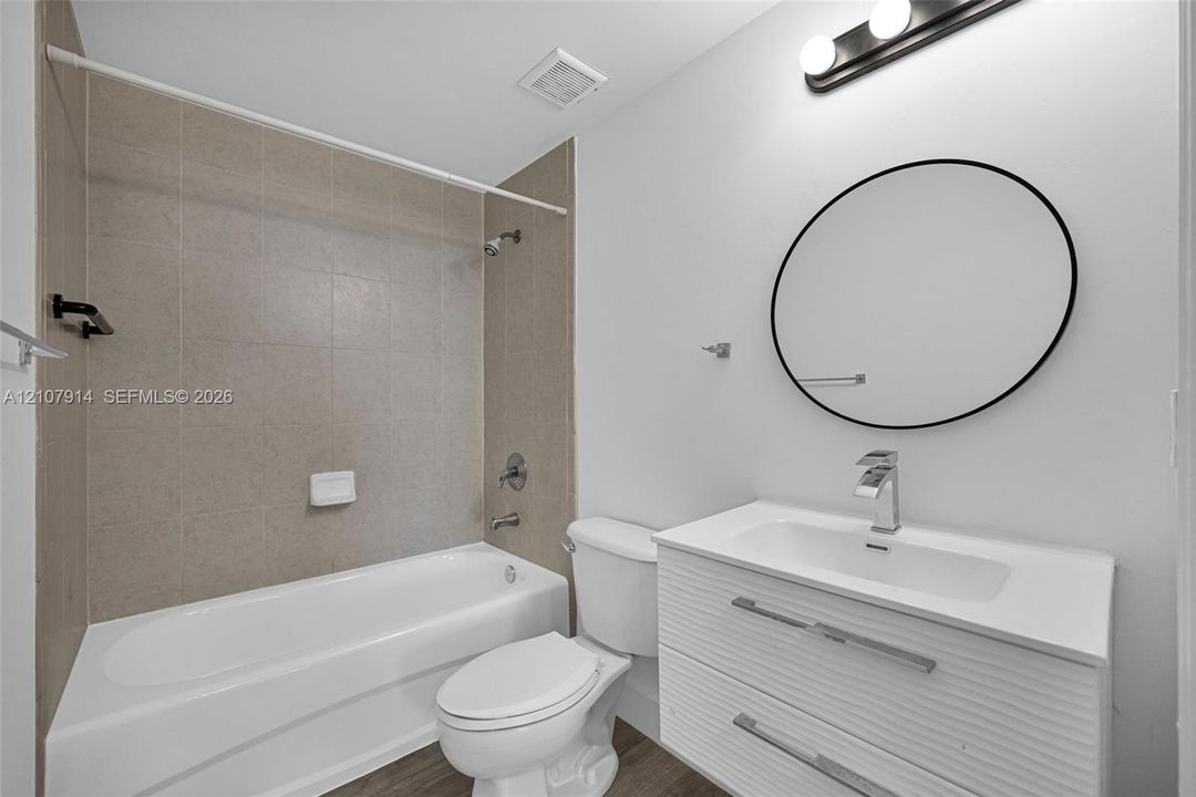 Modern bathroom featuring a floating vanity with a white countertop, circular wall-mounted mirror, three-light fixture, and bathtub with neutral wall tile