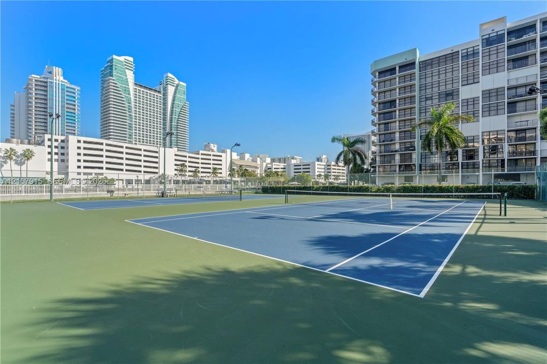 Dual hard-court tennis facility featuring blue and green surfacing, perimeter chain-link fencing, and pole-mounted lighting