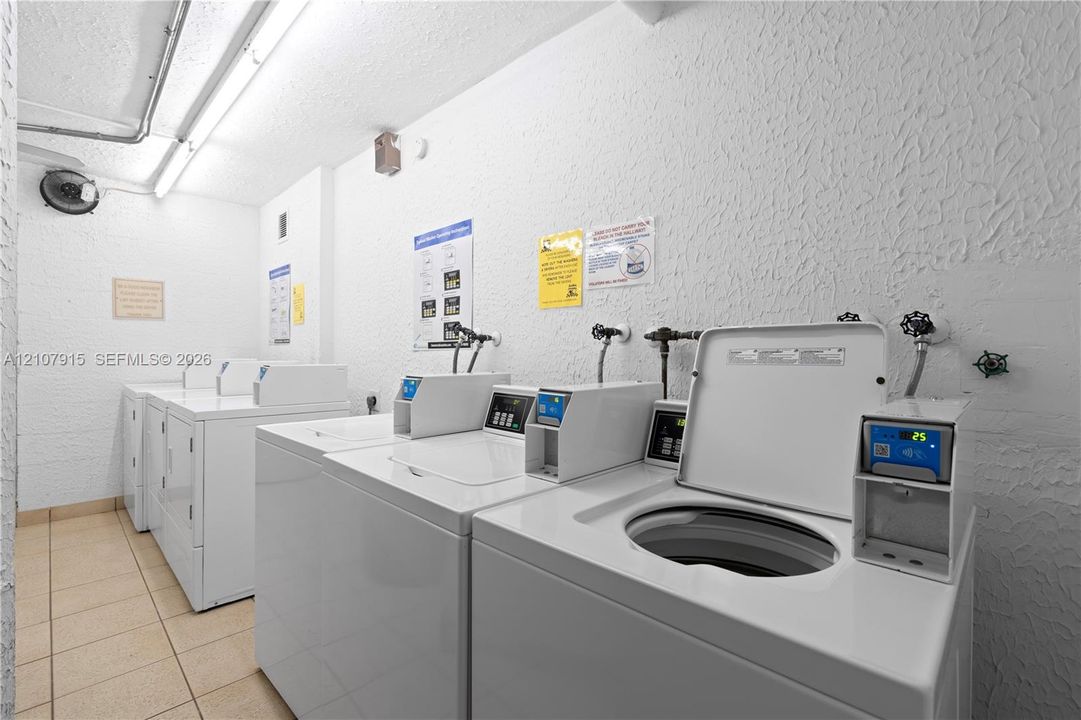Laundry room equipped with multiple top-loading washing machines and neutral tile flooring