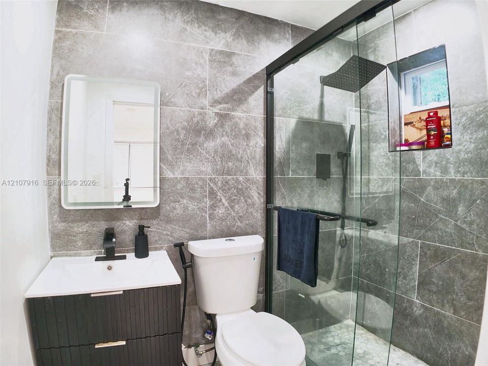 Bathroom featuring large-format grey stone-finish tile, dark wood-finish vanity with a white rectangular sink, matte black hardware, and a frameless glass-enclosed shower with a rainfall showerhead