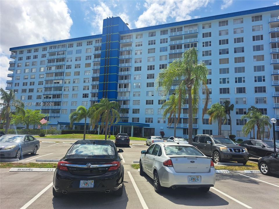 Multi-story residential building featuring a blue exterior facade, private balconies, and dedicated parking area with mature palm trees