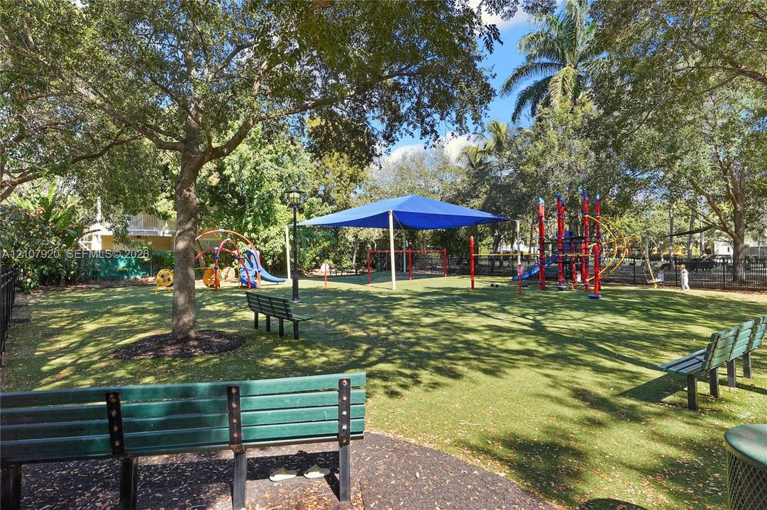 Recreational outdoor space featuring permanent playground structures, a large shade canopy, mature shade trees, and built-in metal bench seating