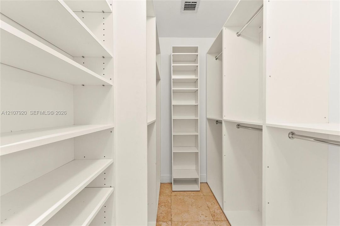 Walk-in closet featuring white custom built-in shelving, adjustable storage units, metal garment rods, and stone-tile flooring
