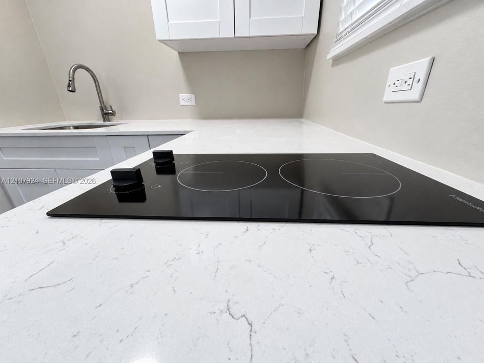 Kitchen area featuring light-toned stone-finish countertops, a black induction cooktop, white shaker-style cabinetry, and an undermount stainless steel sink with a gooseneck faucet