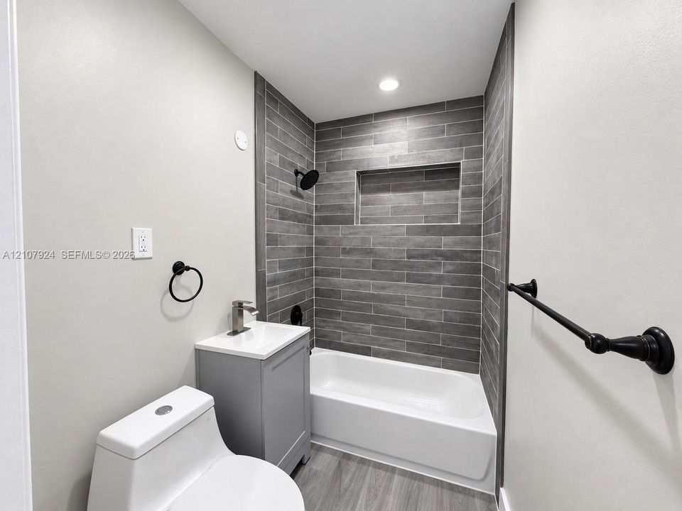 Bathroom featuring gray horizontal tile shower surround with integrated wall niche, wood-finish flooring, vanity with countertop basin, and matte black hardware