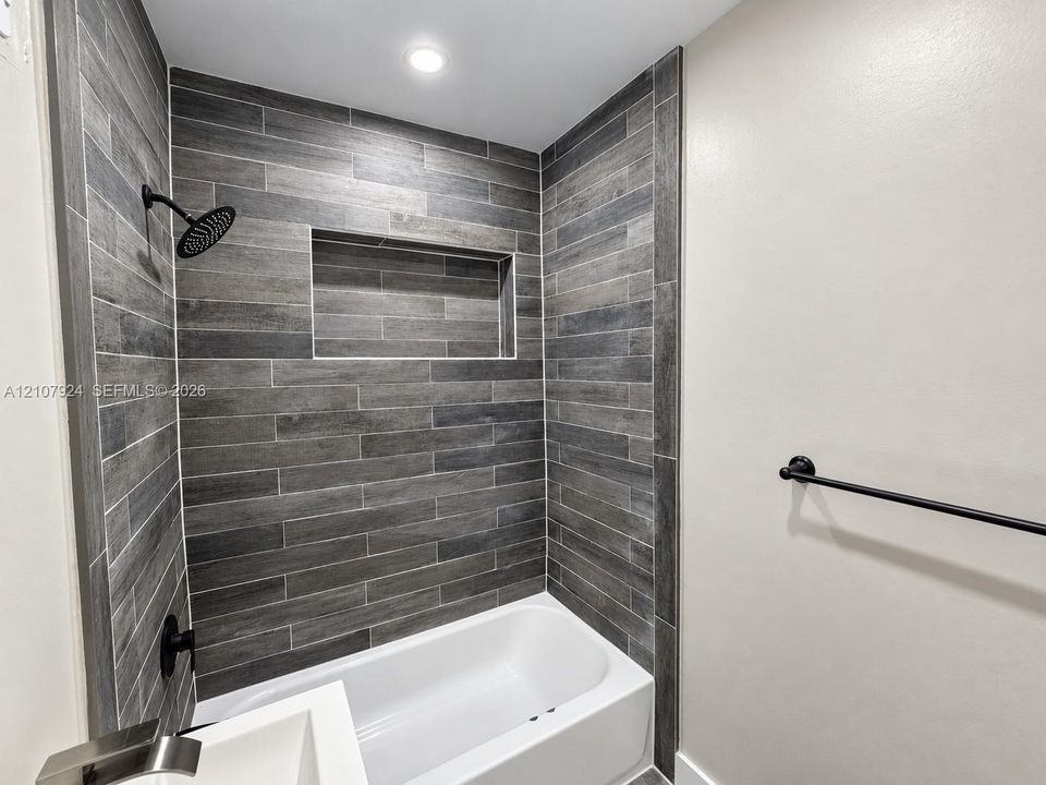 Shower enclosure featuring grey horizontal tile surround, recessed wall niche, matte black rainfall showerhead, and a white soaking tub