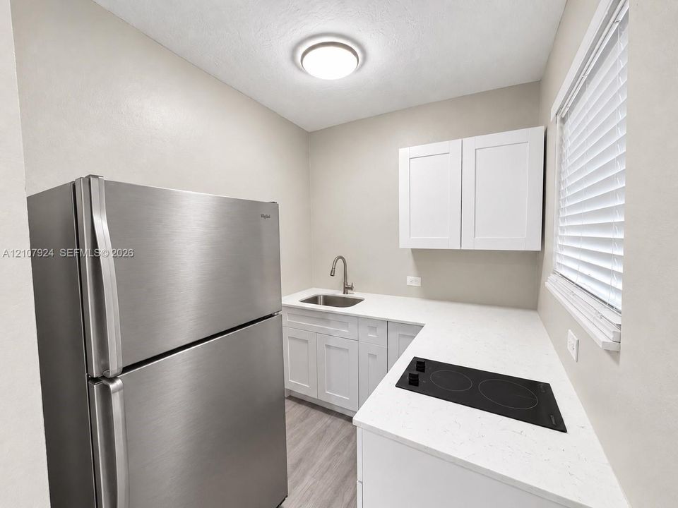 Kitchenette featuring white Shaker-style cabinetry, white stone-finish countertops, an integrated stainless steel refrigerator, an inset stainless steel sink with a high-arc faucet, and a black glass cooktop