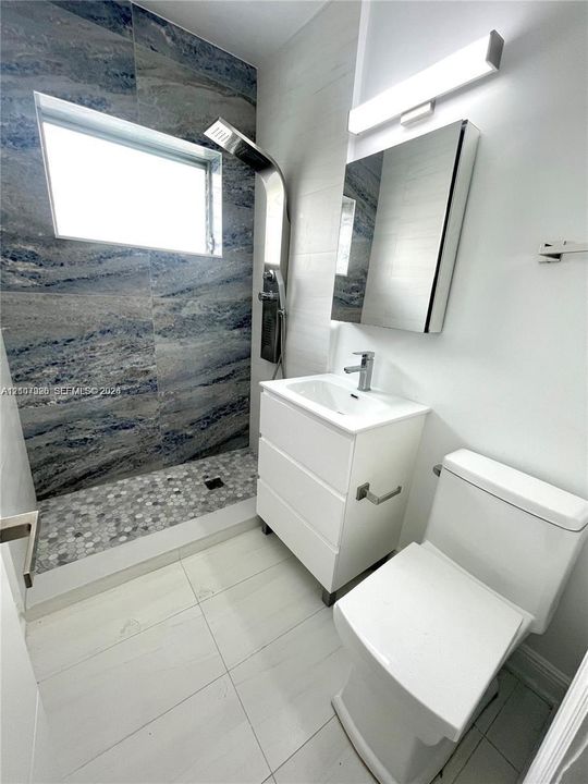 Bathroom featuring a walk-in shower with stone-look wall tiling and a pebble-tiled floor, a wall-mounted shower panel system, a white vanity with an integrated sink, and a mirrored medicine cabinet