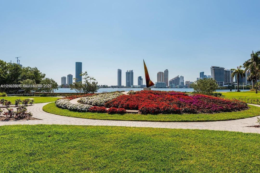 Manicured lawn featuring a circular stone paver path, expansive floral landscaping, decorative sculpture, and water frontage with a cityscape backdrop