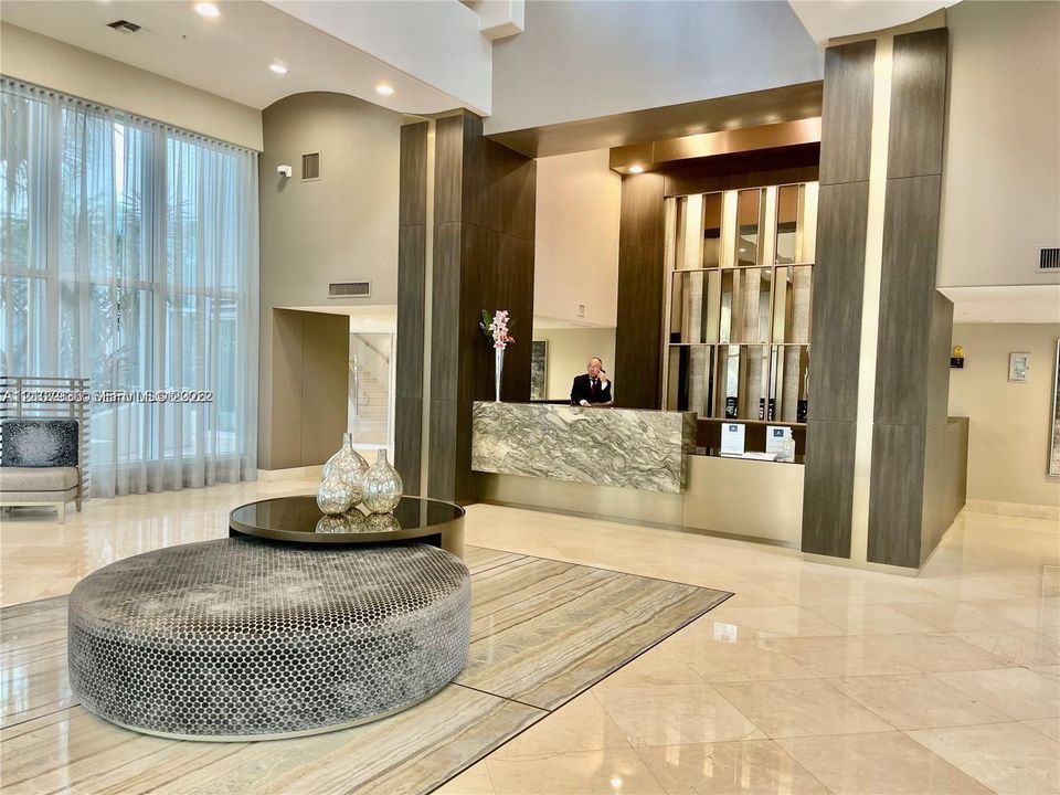Double-height lobby featuring floor-to-ceiling windows, polished stone flooring, and a decorative stone-front reception desk accented with metallic paneling and dark wood-finish columns