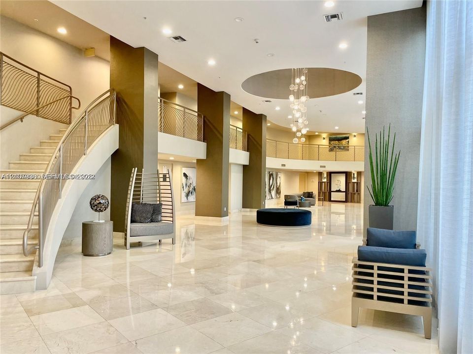 Grand lobby featuring polished stone flooring, a sweeping staircase with custom metal railing, multi-level mezzanine, and a recessed ceiling with a contemporary pendant chandelier