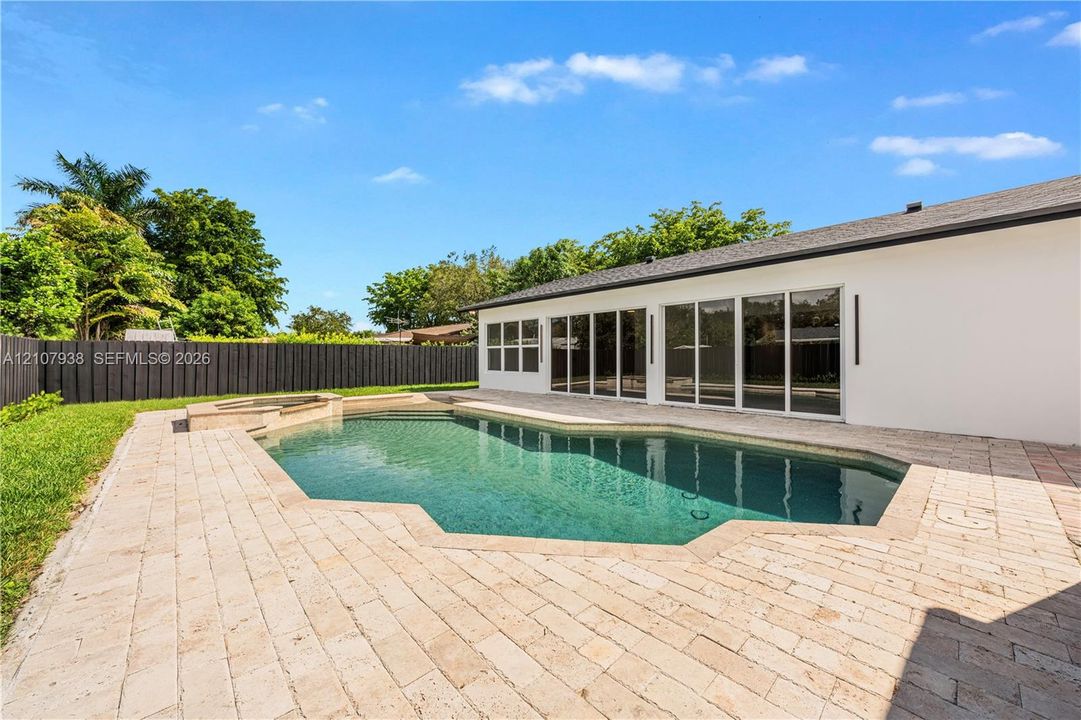 Custom-shaped in-ground pool featuring a built-in spa, expansive stone paver deck, white exterior wall, and floor-to-ceiling glass sliding doors