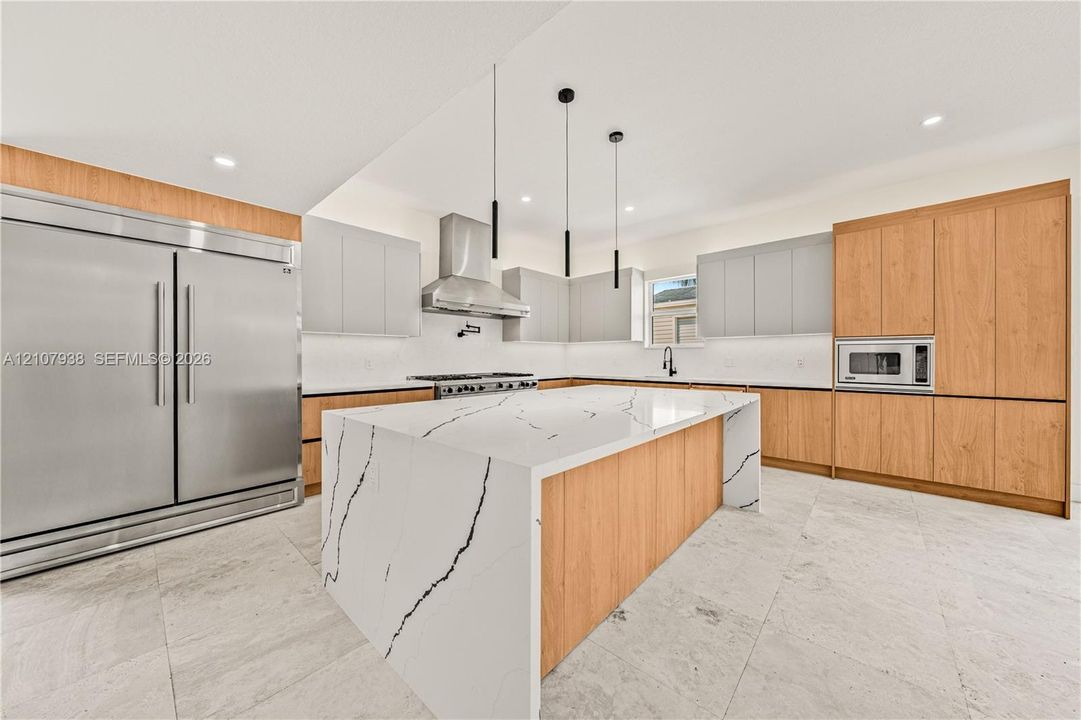 Kitchen featuring a large island with a stone-finish waterfall countertop, wood-finish cabinetry, stainless steel professional-grade appliances, under-cabinet lighting, and stone flooring