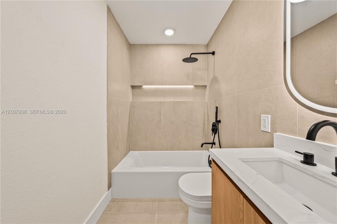 Bathroom featuring a vanity with a light wood-finish cabinet, stone-surface countertop, and matte black fixtures, paired with a shower-tub combination featuring recessed wall niche lighting and floor-to-ceiling tile work