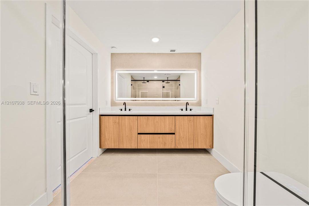 Floating wood-finish double vanity with stone countertop, dual undermount sinks, matte black widespread faucets, and a large integrated LED-backlit mirror