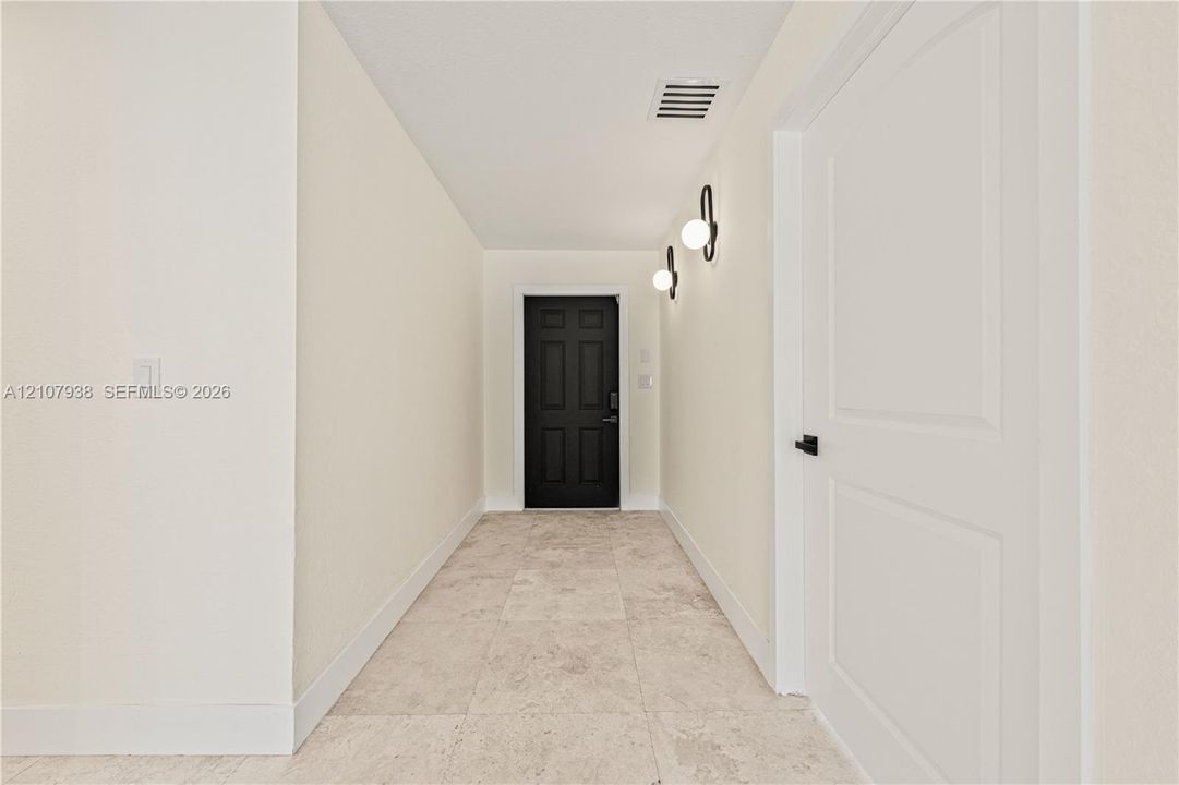 Neutral-toned stone-finish flooring, six-panel black door, dual globe-style wall-mounted light fixtures, and crisp white interior doors with matte black hardware