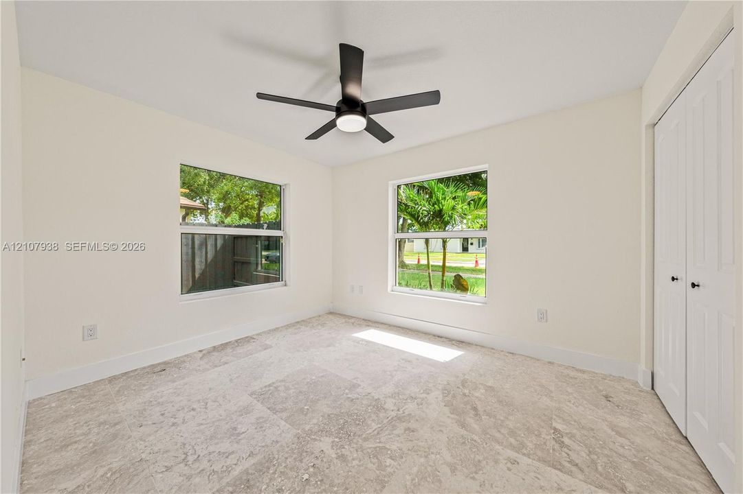 Neutral-toned tile flooring, dual windows with natural light, a ceiling-mounted fan with integrated lighting, and white bi-fold closet doors