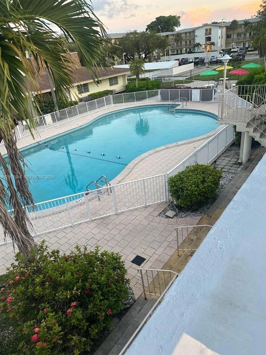 In-ground swimming pool surrounded by a paver deck, white metal perimeter fencing, and established tropical landscaping