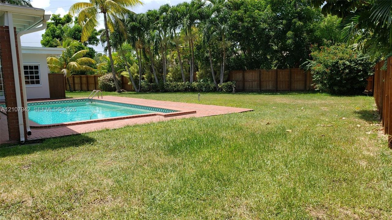 Spacious backyard featuring an in-ground swimming pool, brick paver decking, privacy fencing, and mature palm trees