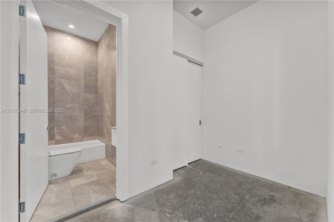Interior space featuring polished concrete flooring and a sliding door, revealing a bathroom finished with large-format tile flooring and wall surfacing, a white bathtub, and a wall-mounted toilet
