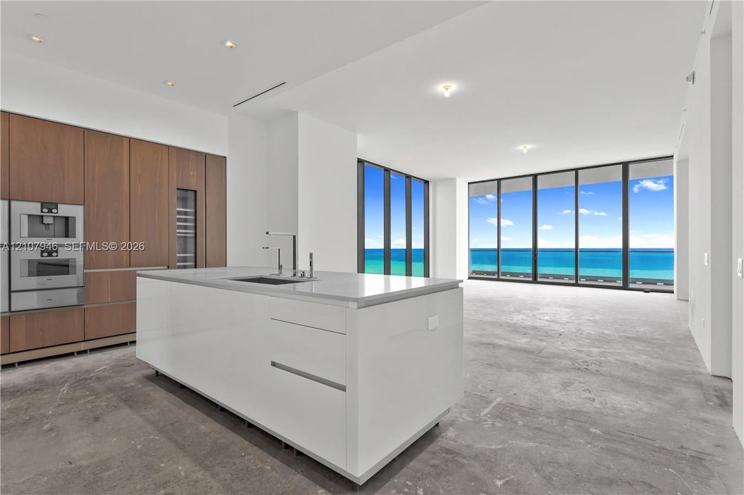 Kitchen featuring full-height wood-finish cabinetry, built-in stainless steel appliances, stone-surface center island with integrated sink, and floor-to-ceiling windows with ocean backdrop