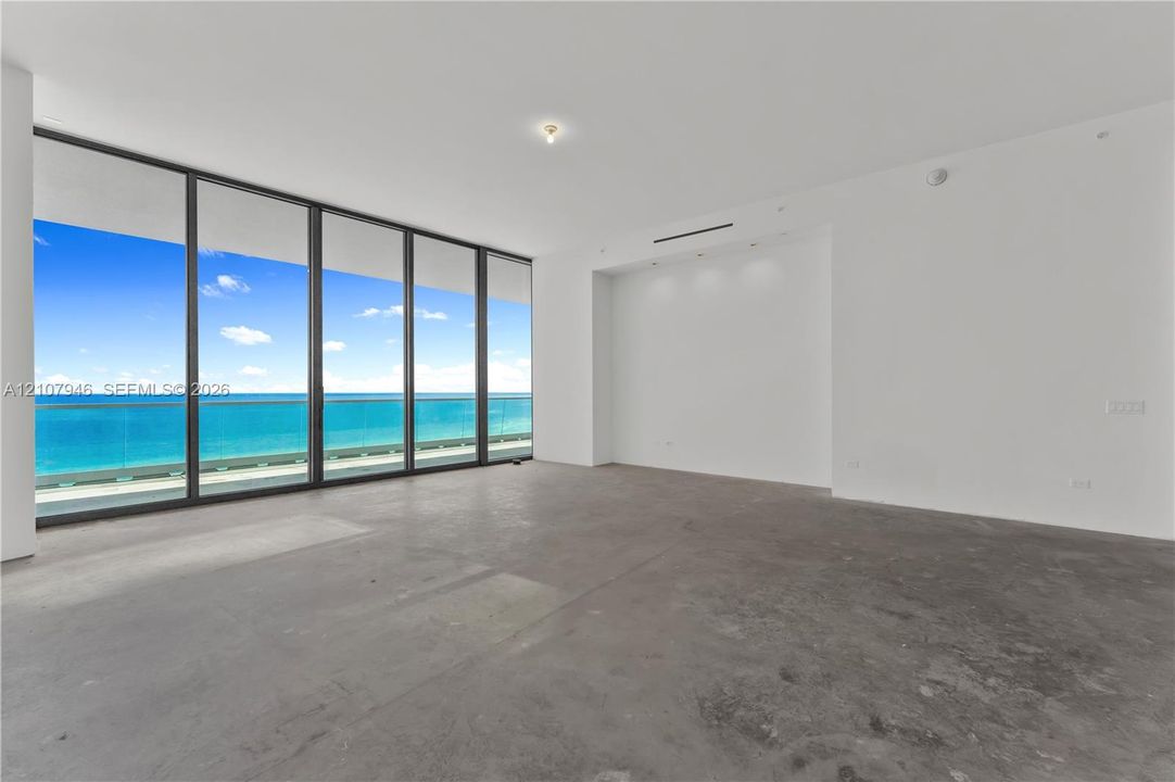 Floor-to-ceiling windows, full-height glass sliding doors, private glass-railed balcony, polished concrete flooring, and ocean backdrop