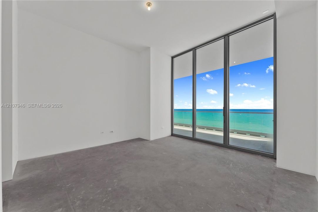 Floor-to-ceiling windows featuring ocean horizon scenery, concrete flooring, and an attached balcony with a glass railing