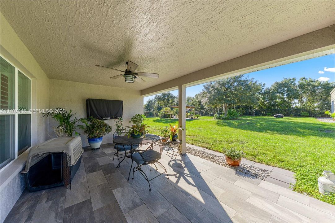 Covered patio featuring ceramic tile flooring, a ceiling-mounted fan, and a built-in television wall mount, overlooking a spacious lawn with mature tree line