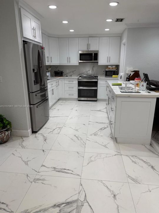 Kitchen featuring white shaker-style cabinetry, stainless steel appliances, a central island with a stone-finish countertop, and large-format tile flooring with a veined pattern