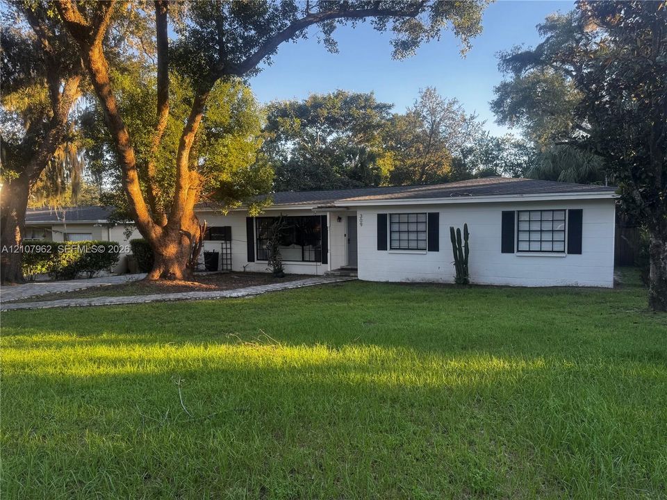 Single-story exterior featuring a light-colored masonry façade, dark-framed window shutters, and a sprawling front lawn with mature canopy trees