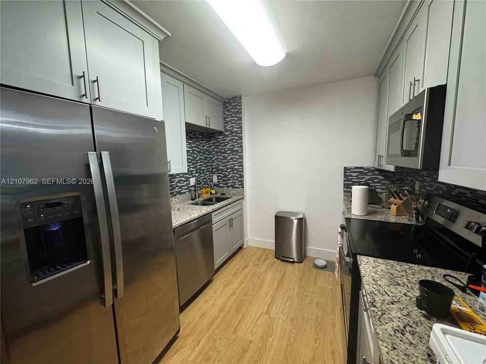 Kitchen featuring light gray shaker-style cabinetry, speckled stone countertops, a decorative mosaic backsplash, stainless steel appliances, and wood-finish flooring