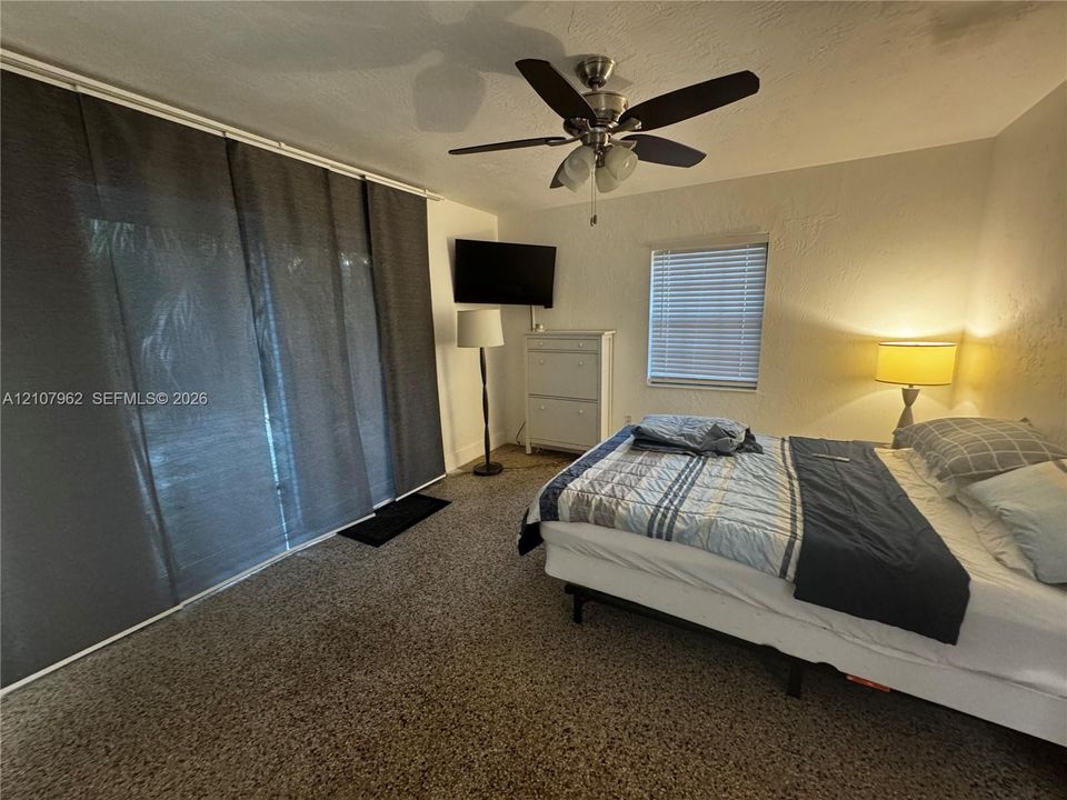 Textured ceiling with a multi-blade ceiling fan and integrated lighting, sliding glass door, and window with horizontal blinds