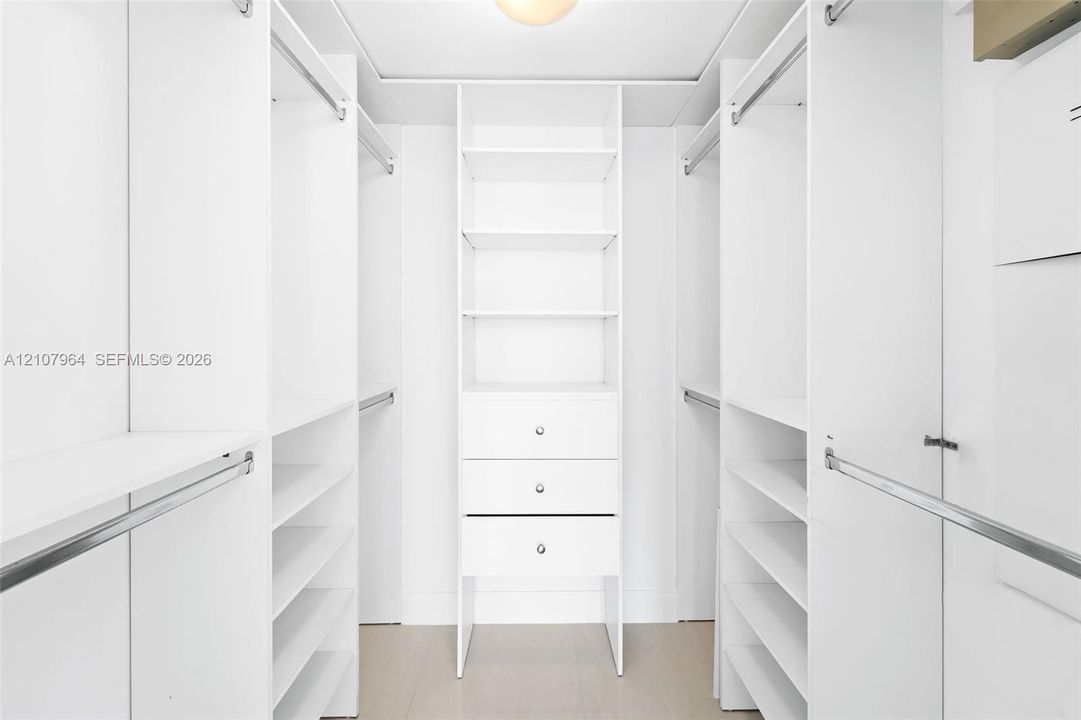 Custom walk-in closet featuring white built-in shelving, triple stacked drawers with hardware, multiple hanging rods, and wood-finish flooring