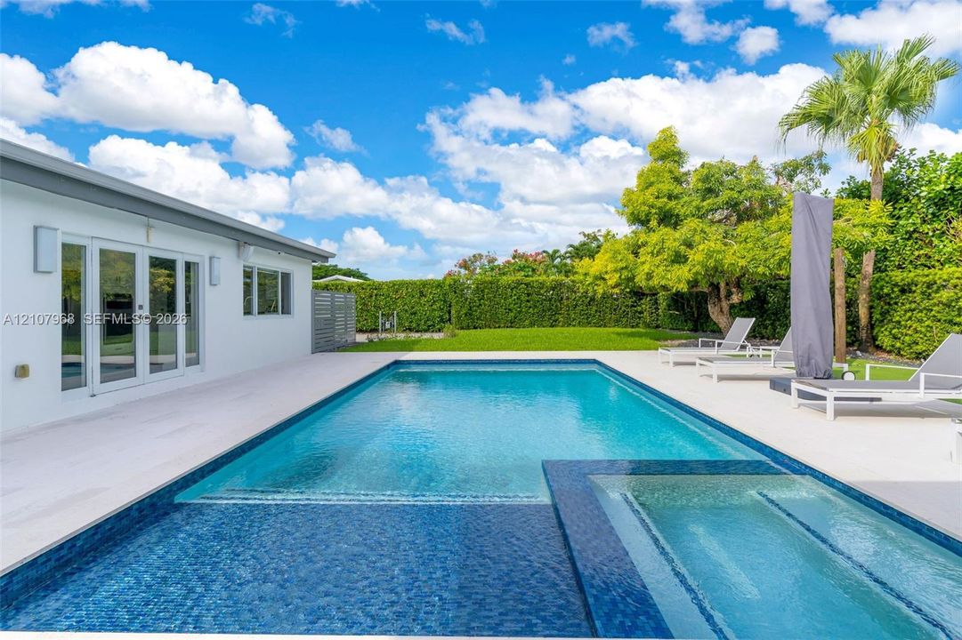 Backyard featuring a custom swimming pool with integrated spa, tile finish, expansive light-toned hardscaping, and privacy hedge