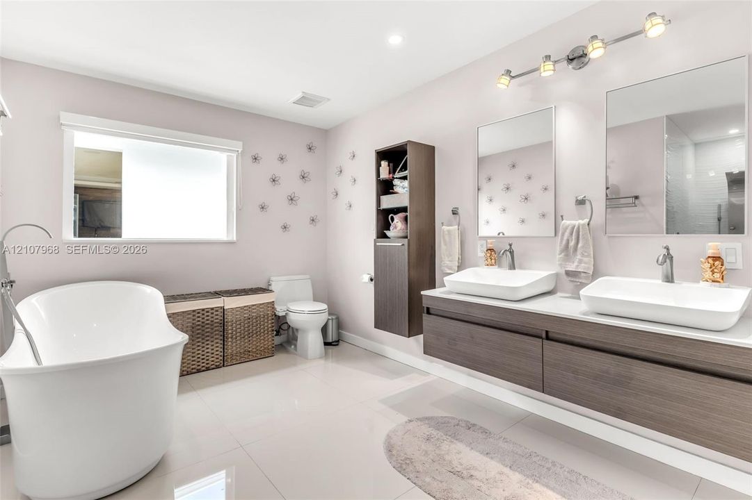 Floating vanity featuring wood-finish cabinetry, dual vessel sinks, and stone-surface countertop, complemented by a freestanding soaking tub, vertical built-in storage cabinet, and rectangular wall-mounted mirrors