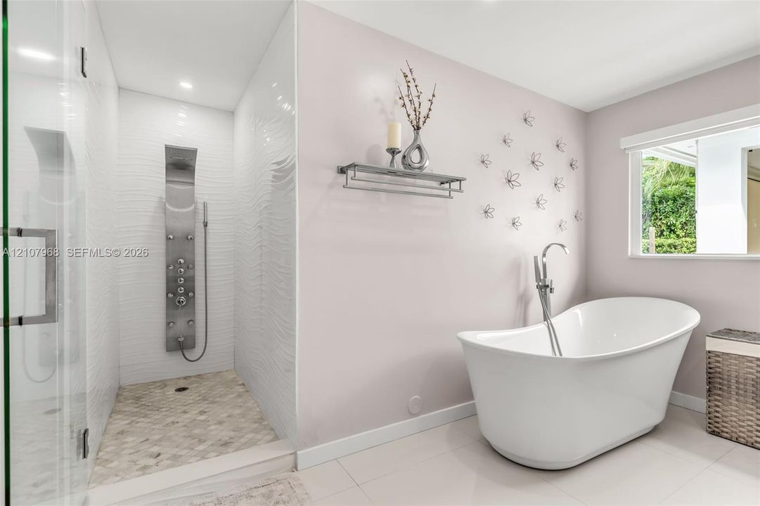 Freestanding soaking tub with a floor-mounted faucet, walk-in shower featuring textured wall tile, stainless steel rainfall shower panel, and pebble-style shower flooring