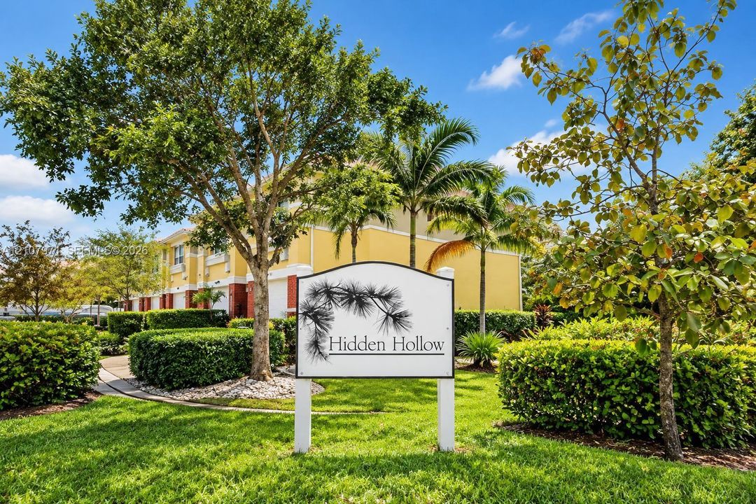 Community entrance sign featuring professional signage, manicured landscaping, mature trees, palm trees, and paved walkways