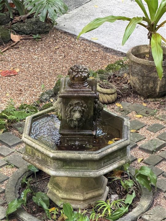 Hexagonal stone water fountain featuring lion head sculptural details, integrated into a landscape setting with paver accents and gravel ground cover
